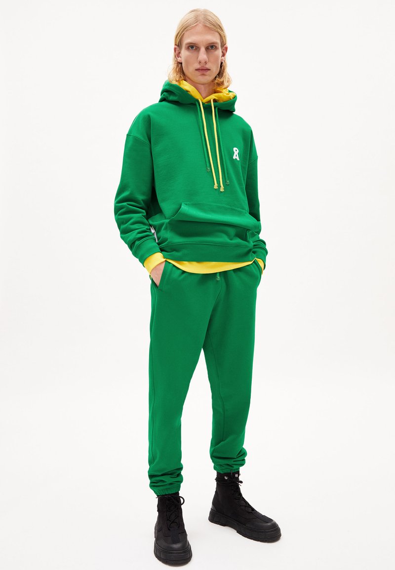 Grüne Kapuzenjacke mit gelben Akzenten und Kordeln, kombiniert mit passenden grünen Jogginghosen. Verfügt über ein weißes Logo und Seitentaschen. Schwarze Stiefel.