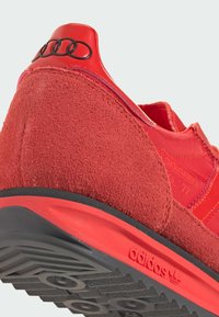 Zapatilla deportiva de ante rojo con detalles de tela, que cuenta con una suela de goma negra. Logotipo grabado en el talón y patrón de rayas distintivo en el lateral.