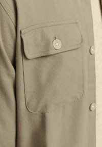 Chemise khaki à boutons avec une poche poitrine à rabats et des boutons beiges, fabriquée à partir d'un tissu texturé avec des détails de coutures visibles.