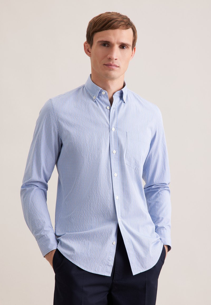 Jeune homme aux cheveux bruns courts portant une chemise bleu clair à boutons et un pantalon foncé, debout les mains dans les poches devant un fond uni.