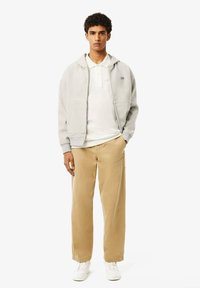 Felpa grigia con zip e tasche laterali, polo bianca sotto, pantaloni a gamba larga beige e sneaker bianche. Modella in piedi contro uno sfondo bianco.