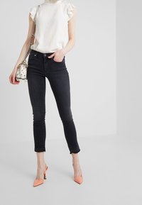 Blusa blanca con volantes, jeans ajustados negros con dobladillo desgastado y tacones en punta melocotón. La modelo sostiene un pequeño bolso transparente con acentos florales.