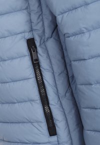 Világoskék puffermellény, steppelt textúrával, fekete cipzárral és anyagfüllel a záráshoz. Puha, szigetelt anyag.