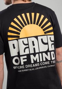 La maglietta nera in cotone presenta una grande grafica del sole giallo con linee radiali sopra il testo in grassetto "PEACE OF MIND" e un testo più piccolo sottostante.