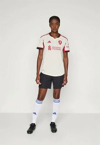 Mujer de pie vestida con camiseta de fútbol blanca del Liverpool FC, pantalones cortos negros, calcetas blancas hasta la rodilla con franjas azules y botas negras, en un fondo sencillo.