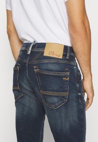 Un jean en denim bleu foncé présente un patch en cuir avec "LTB", des coutures jaunes contrastantes et plusieurs poches. Design ajusté.