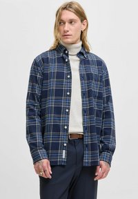 Camisa de franela a cuadros en azul marino con rayas blancas y amarillas, cuello abotonado, bolsillo en el pecho y mangas largas; llevada sobre un cuello de tortuga claro.