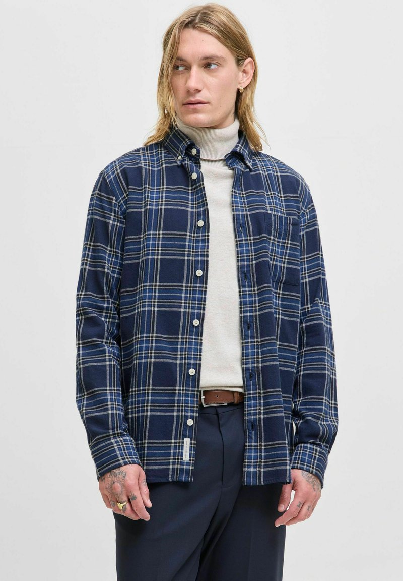 Camisa de franela a cuadros en azul marino con rayas blancas y amarillas, cuello abotonado, bolsillo en el pecho y mangas largas; llevada sobre un cuello de tortuga claro.