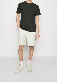 T-shirt a righe nere con collo tondo, abbinata a pantaloni cargo beige chiaro con tasca laterale e patch con logo, indossati con calzini bianchi e scarpe.