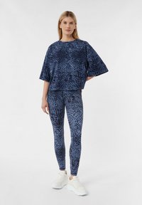 Marineblaues Sportoberteil mit lockerem Schnitt, das ein dezentes Leopardenmuster aufweist, kombiniert mit enganliegenden Leggings im passenden Muster und weißen Sneakers.