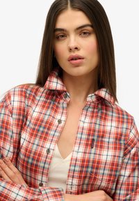 Chemise à carreaux rouge et blanc avec des accents bleus, à boutons et col. Elle est dotée d'une poche sur la poitrine gauche et d'un détail de marque subtil.