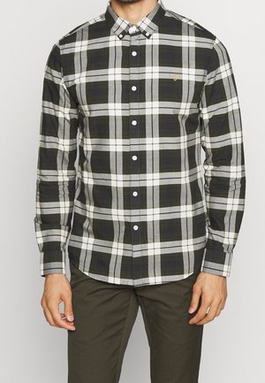 Mand iført en sort, hvid og grå ternet skjorte med lange ærmer og mørke bukser, stående mod en ensfarvet baggrund.