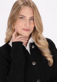 Schwarze Strickjacke mit Zopfmuster, ausgestattet mit silbernen Knopfkreisen. Trägt eine weiße Hemdbluse darunter. Lange blonde Haare sichtbar.