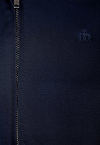 Tela azul oscuro con una cremallera plateada visible en el lado izquierdo y un pequeño logo de corona bordado a la derecha.