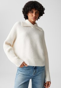 Maglione bianco a coste con colletto ripiegato e maniche larghe, abbinato a jeans di denim blu chiaro. Tessuto morbido con sottili righe orizzontali.