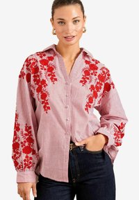 Blusa a righe rosse e bianche con ricami floreali sulle maniche, colletto e chiusura con bottoni. Tessuto morbido con una vestibilità rilassata.