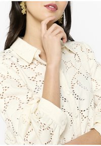 Camicia bianca in cotone con intricati motivi a taglio, colletto e chiusura con bottoni, abbinata a orecchini floreali dorati.