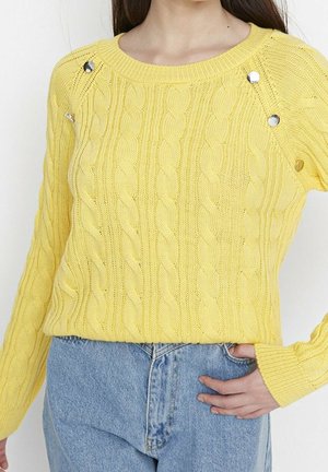 Femme portant un pull jaune torsadé avec des boutons argentés sur les épaules et un jean taille haute bleu clair, main dans la poche.