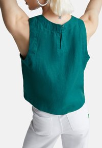 Tyrkysový lněný top bez rukávů s kulatým výstřihem, zapínáním na knoflík vzadu a mírně zvýšeným spodním lemem. V kombinaci s bílými džíny, zobrazený zezadu.