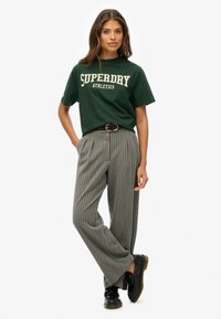 Camiseta de algodón verde con el logo "SUPERDRY ATHLETICS", combinada con pantalones de pierna ancha rayados en gris y zapatos negros con cordones.