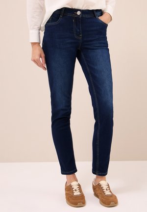Woman wearing dark blue slim-fit jeans and brown suede sneakers, standing with one hand in pocket against neutral background.
