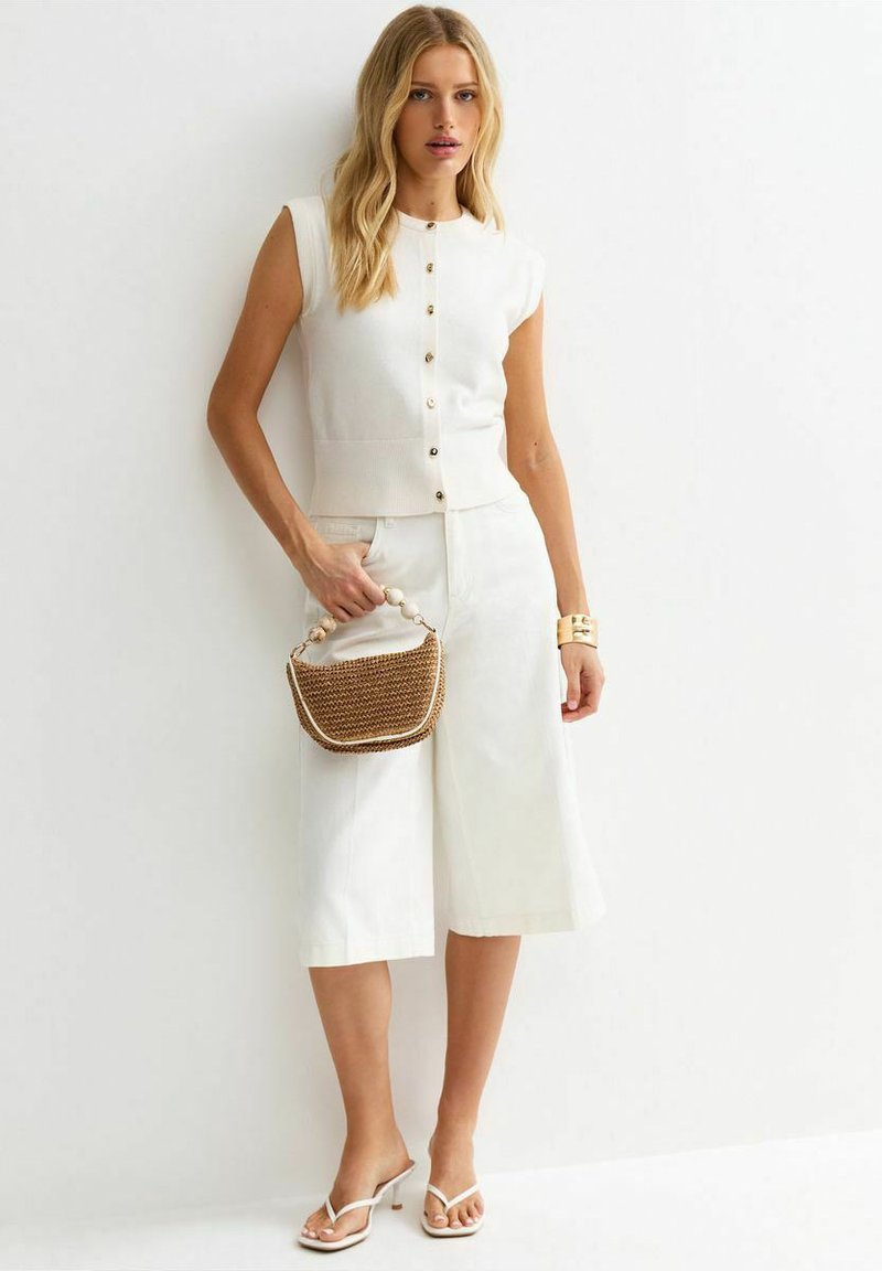 Woman wearing sleeveless white buttoned top and white wide-leg cropped pants, holding a small woven handbag with bead handle.