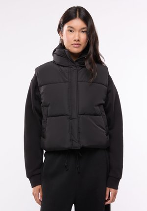 Giovane donna con lunghi capelli scuri che indossa un gilet imbottito nero sopra una felpa nera con cappuccio a maniche lunghe e pantaloni neri, in piedi davanti a uno sfondo bianco.