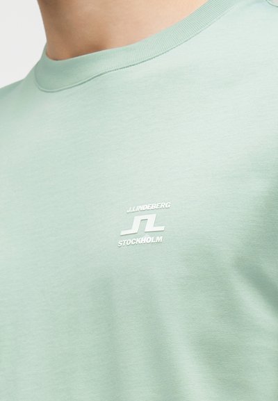 Κοντινό πλάνο σε φωτεινό πράσινο t-shirt με στρογγυλή λαιμόκοψη και λευκό λογότυπο J.LINDEBERG Stockholm στην αριστερή πλευρά του στήθους.