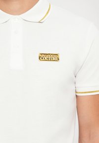 Hvit polo skjorte laget av teksturert stoff, med krage i gullstriper og en gull logo-plakett på brystet.