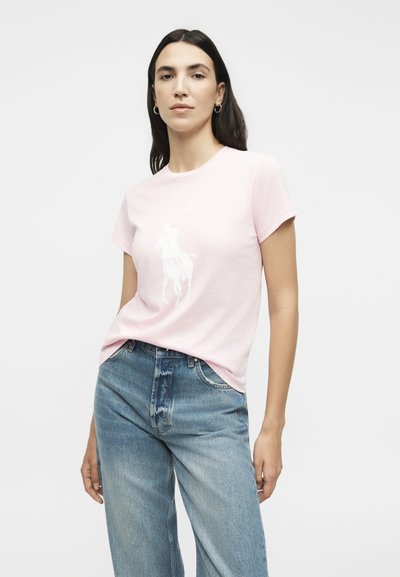 Γυναίκα με μακριά σκούρα μαλλιά που φοράει ένα απαλό ροζ μπλουζάκι με λευκό γραφικό παίκτη πόλο και ανοιχτά μπλε τζιν ψηλόμεσα παντελόνια.