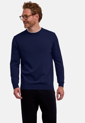 Hombre con cabello rizado y gafas, vistiendo un suéter azul marino de manga larga y pantalones negros, mirando hacia la izquierda y sonriendo.