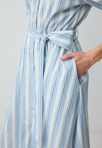 Abito a righe blu e bianche con vita legata, chiusura a bottoni davanti e tasca laterale. Il materiale appare leggero e morbido.