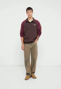 Μπορντό και σκούρο καφέ πουλόβερ με γιακά και λογότυπο· συνδυασμένο με ανοιχτό καφέ κωνικά παντελόνια και καφέ αθλητικά παπούτσια. Απλός, καθημερινός σχεδιασμός.