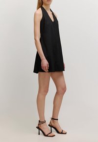 Robe noire à col licou avec décolleté en V et ourlet à hauteur des genoux. Fabriquée en tissu léger ; texture lisse et design simple. Portée avec des talons noirs à brides.