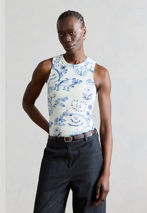 Femme aux cheveux tressés portant un haut blanc sans manches avec des motifs floraux bleus et un pantalon taille haute foncé avec une ceinture marron.