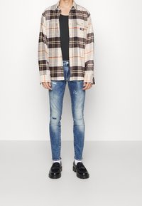 Camisa de cuadros en tonos beige, marrón y naranja, combinada con jeans azules desgastados y mocasines negros. Cuenta con mangas largas y bolsillos delanteros.