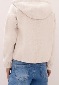 Jasna beżowa kurtka polarowa z kapturem, o fakturowanej powierzchni. Elastyczne mankiety i krótki dół. Noszona z niebieskimi jeansami.