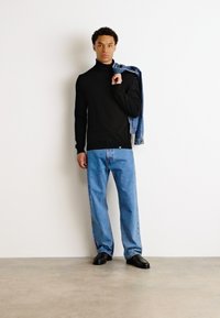 Μαύρο πουλόβερ με ζιβάγκο, χαλαρές μπλε τζιν τζάκετ, και μαύρα παπούτσια. Ένα τζιν τζάκετ είναι πεταμένο στον ώμο. Ουδέτερο φόντο.