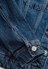 Gros plan d'une manche de veste en jean bleu avec des coutures visibles et un bouton métallique étiqueté "Next Jeans Since 1982."