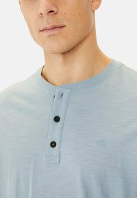 Lyseblå, kortærmet henley skjorte lavet af blødt stof, der har tre sorte knapper og et lille broderet logo på brystet.