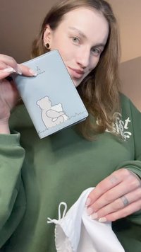 Lyseblå pung med præget bjørnemotiv, der har en glat tekstur og syninger ved kanterne. En hvid snørepose holdes i den anden hånd.