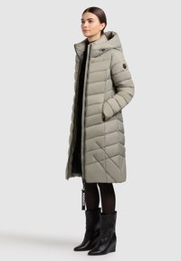 Pitkä, vaaleanharmaa toppatakki hupulla, jossa timanttikuvioinen tikkaus, vetoketju edessä ja sivutaskut. Päällä mustan mekon ja mustien saappaiden päällä.