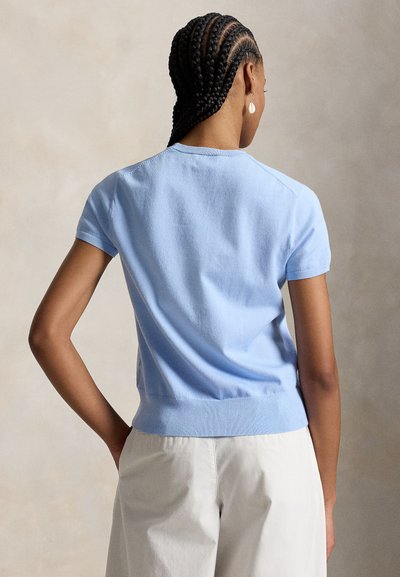 Kvinne med flette, iført lys blå kortermet genser og hvite bukser, sett bakfra mot en nøytral bakgrunn.