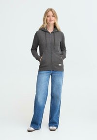 Felpa grigia con zip e cappuccio, con lacci, tasche frontali e un patch logo, abbinata a jeans a zampa d'elefante blu chiaro e scarpe grigie.