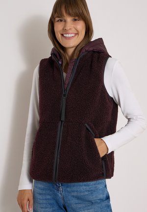 Femme souriante porte une veste polaire zippée violet foncé avec capuche sur une chemise blanche à manches longues et un jean bleu, main dans la poche.