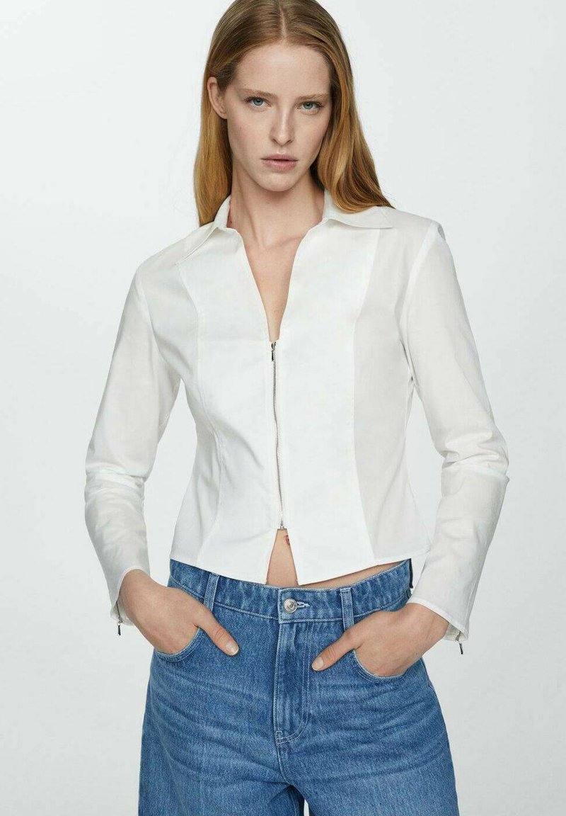 Weiße Zip-Jacke mit Kragen, tailliertem Schnitt und langen Ärmeln. Getragen über hochgeschnittenen blauen Jeans. Das Material wirkt glatt und leicht.