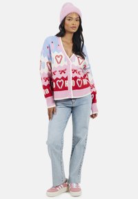 Cardigan lavorato a maglia colorato con motivi rosa, rosso e blu, decorato con cuori e fiocchi; chiusura a bottoni, maniche lunghe e tessuto texture. Abbinato a jeans chiari e scarpe da ginnastica rosa.