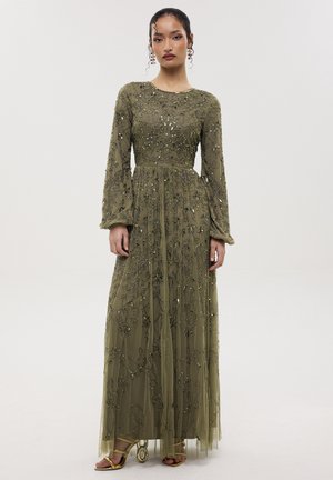 Femme portant une robe longue vert olive avec un travail de perles complexe et de longues manches transparentes, associée à des talons ouverts dorés.