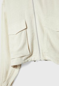 Gros plan d'une veste couleur crème avec fermeture éclair à l'avant et deux poches à rabat sur la poitrine, en tissu texturé avec poignets froncés.