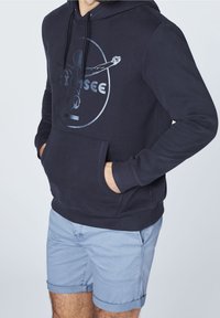 Marinblå hoodie med en framficka och justerbar dragsko på huvan. Har en grafisk tryck och ribbade ärmslut. Matchas med ljusblå shorts.